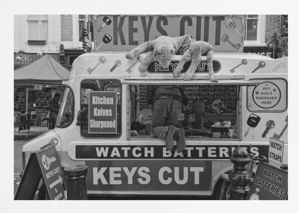 A quirky black and white photograph of a mobile key-cutting van on a city street. A man in jeans is seen from behind, kneeling inside the service window. On the roof, a large stuffed tiger lies stretched out, peering down. The van is covered in signs advertising 'KEYS CUT', 'WATCH BATTERIES', and 'Kitchen Knives Sharpened'. Print