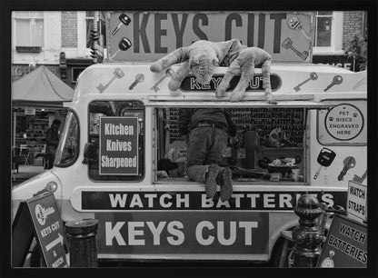 A quirky black and white photograph of a mobile key-cutting van on a city street. A man in jeans is seen from behind, kneeling inside the service window. On the roof, a large stuffed tiger lies stretched out, peering down. The van is covered in signs advertising 'KEYS CUT', 'WATCH BATTERIES', and 'Kitchen Knives Sharpened'. Print
