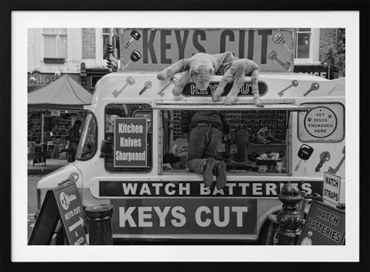 A quirky black and white photograph of a mobile key-cutting van on a city street. A man in jeans is seen from behind, kneeling inside the service window. On the roof, a large stuffed tiger lies stretched out, peering down. The van is covered in signs advertising 'KEYS CUT', 'WATCH BATTERIES', and 'Kitchen Knives Sharpened'. Print