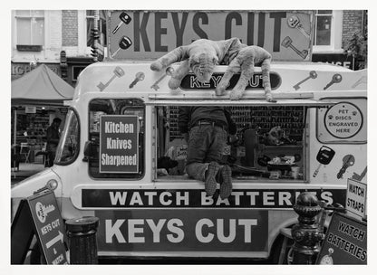 A quirky black and white photograph of a mobile key-cutting van on a city street. A man in jeans is seen from behind, kneeling inside the service window. On the roof, a large stuffed tiger lies stretched out, peering down. The van is covered in signs advertising 'KEYS CUT', 'WATCH BATTERIES', and 'Kitchen Knives Sharpened'. Print