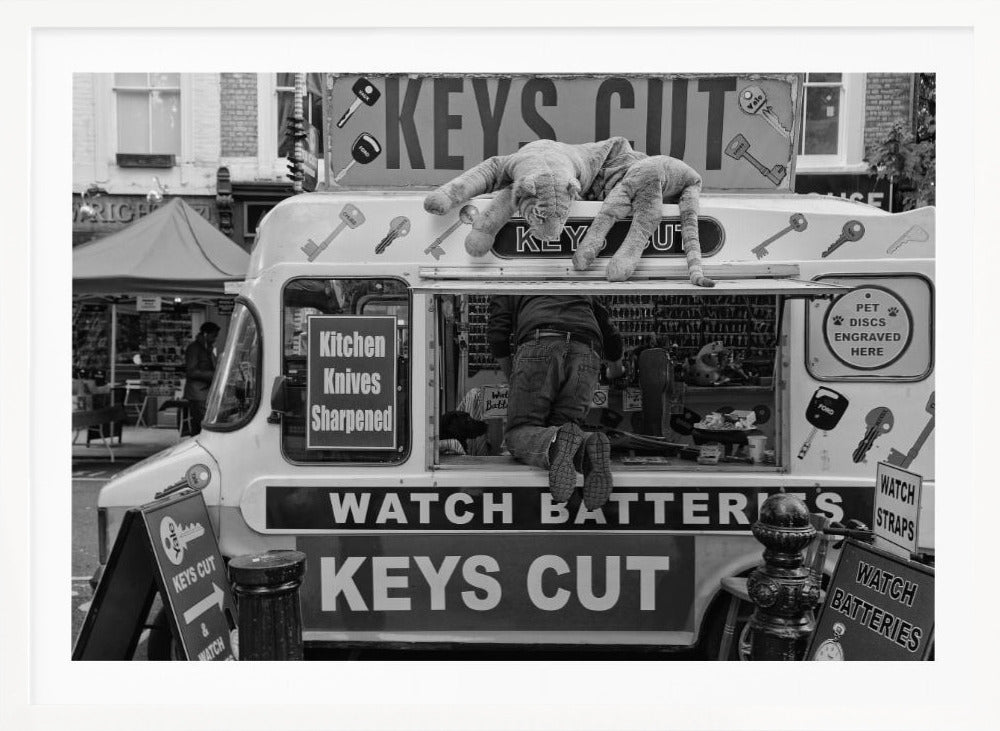 A quirky black and white photograph of a mobile key-cutting van on a city street. A man in jeans is seen from behind, kneeling inside the service window. On the roof, a large stuffed tiger lies stretched out, peering down. The van is covered in signs advertising 'KEYS CUT', 'WATCH BATTERIES', and 'Kitchen Knives Sharpened'. Print