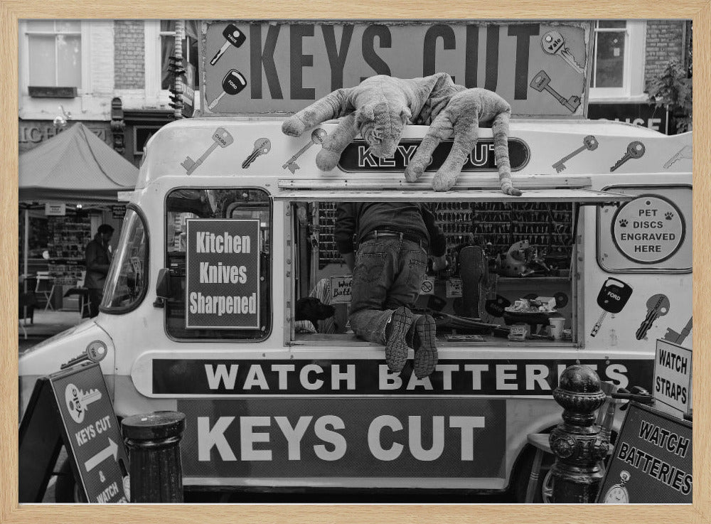 A quirky black and white photograph of a mobile key-cutting van on a city street. A man in jeans is seen from behind, kneeling inside the service window. On the roof, a large stuffed tiger lies stretched out, peering down. The van is covered in signs advertising 'KEYS CUT', 'WATCH BATTERIES', and 'Kitchen Knives Sharpened'. Print