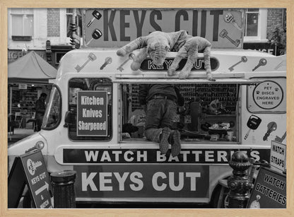 A quirky black and white photograph of a mobile key-cutting van on a city street. A man in jeans is seen from behind, kneeling inside the service window. On the roof, a large stuffed tiger lies stretched out, peering down. The van is covered in signs advertising 'KEYS CUT', 'WATCH BATTERIES', and 'Kitchen Knives Sharpened'. Print