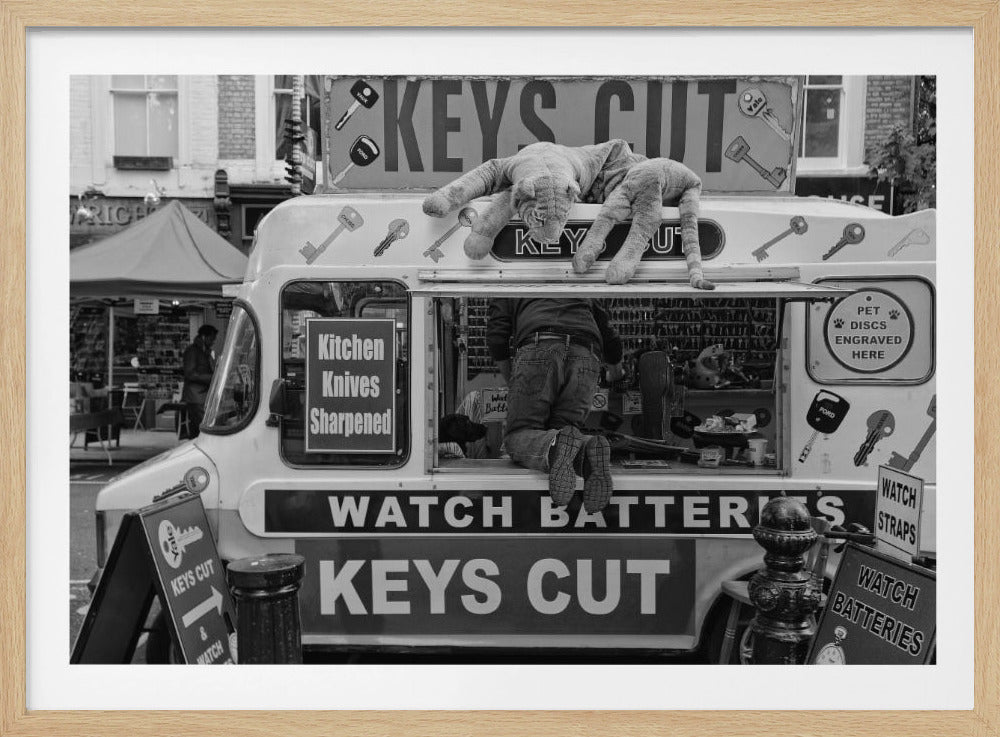 A quirky black and white photograph of a mobile key-cutting van on a city street. A man in jeans is seen from behind, kneeling inside the service window. On the roof, a large stuffed tiger lies stretched out, peering down. The van is covered in signs advertising 'KEYS CUT', 'WATCH BATTERIES', and 'Kitchen Knives Sharpened'. Print