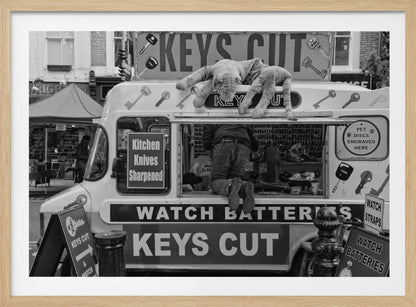 A quirky black and white photograph of a mobile key-cutting van on a city street. A man in jeans is seen from behind, kneeling inside the service window. On the roof, a large stuffed tiger lies stretched out, peering down. The van is covered in signs advertising 'KEYS CUT', 'WATCH BATTERIES', and 'Kitchen Knives Sharpened'. Print