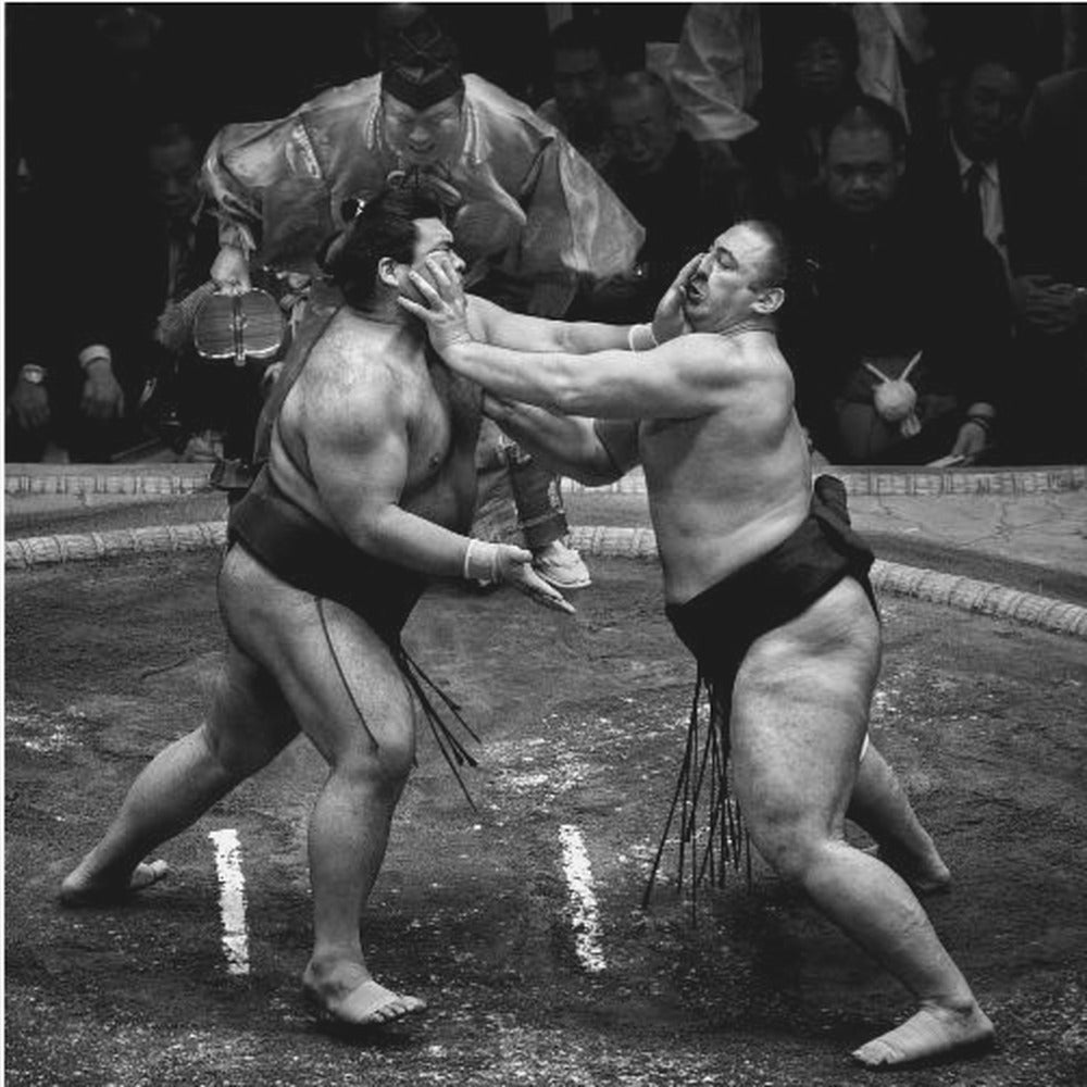A dynamic black and white action photograph of a sumo wrestling match. Two large wrestlers are in the middle of a clay ring, one pushing forcefully against the other's face. A referee in traditional robes stands behind them, and spectators watch from the background. The entire scene is captured within a light wood frame. Artwork