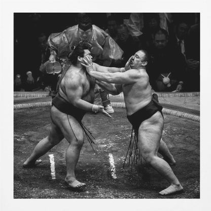 A dynamic black and white action photograph of a sumo wrestling match. Two large wrestlers are in the middle of a clay ring, one pushing forcefully against the other's face. A referee in traditional robes stands behind them, and spectators watch from the background. The entire scene is captured within a light wood frame. Artwork
