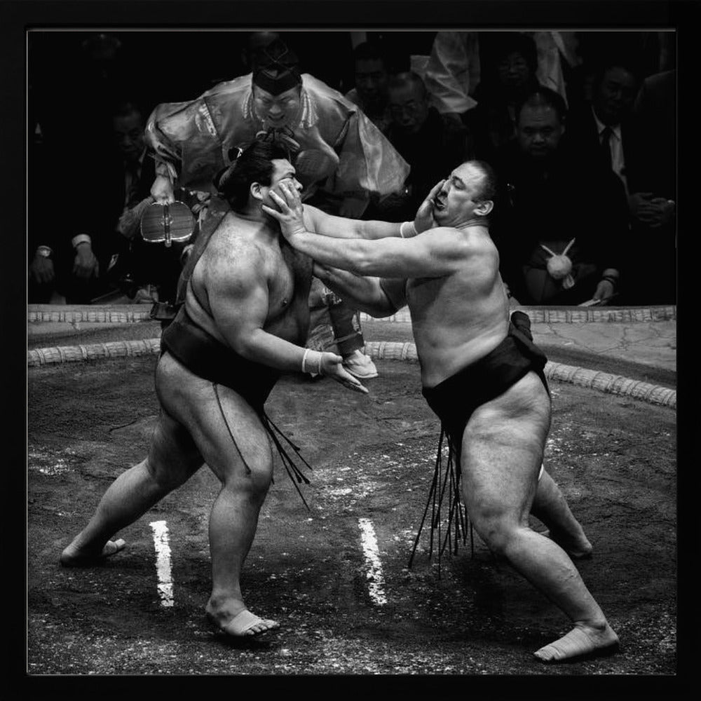 A dynamic black and white action photograph of a sumo wrestling match. Two large wrestlers are in the middle of a clay ring, one pushing forcefully against the other's face. A referee in traditional robes stands behind them, and spectators watch from the background. The entire scene is captured within a light wood frame. Artwork