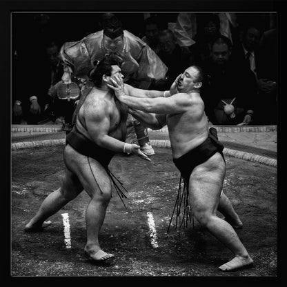 A dynamic black and white action photograph of a sumo wrestling match. Two large wrestlers are in the middle of a clay ring, one pushing forcefully against the other's face. A referee in traditional robes stands behind them, and spectators watch from the background. The entire scene is captured within a light wood frame. Artwork