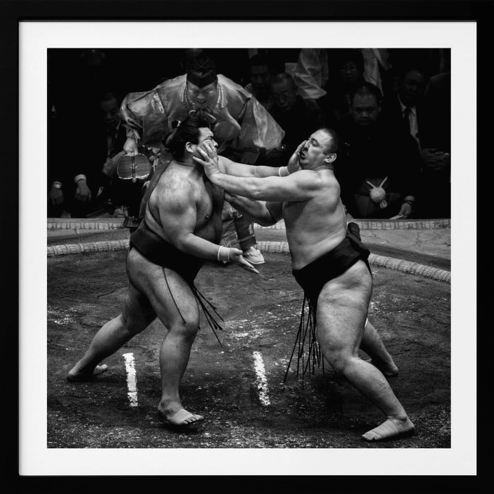 A dynamic black and white action photograph of a sumo wrestling match. Two large wrestlers are in the middle of a clay ring, one pushing forcefully against the other's face. A referee in traditional robes stands behind them, and spectators watch from the background. The entire scene is captured within a light wood frame. Artwork