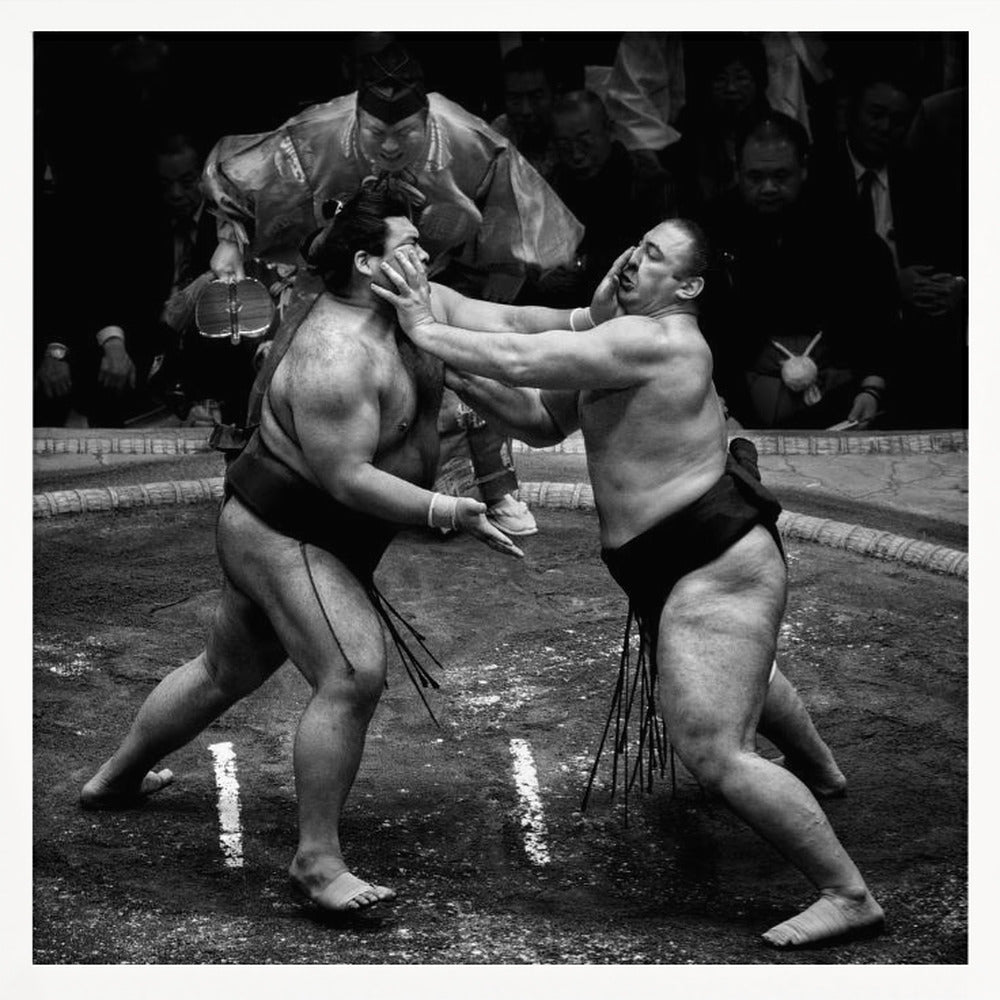 A dynamic black and white action photograph of a sumo wrestling match. Two large wrestlers are in the middle of a clay ring, one pushing forcefully against the other's face. A referee in traditional robes stands behind them, and spectators watch from the background. The entire scene is captured within a light wood frame. Artwork