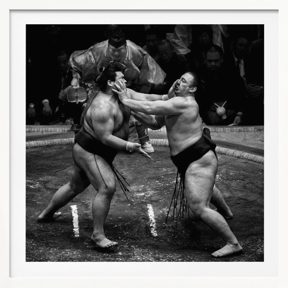 A dynamic black and white action photograph of a sumo wrestling match. Two large wrestlers are in the middle of a clay ring, one pushing forcefully against the other's face. A referee in traditional robes stands behind them, and spectators watch from the background. The entire scene is captured within a light wood frame. Artwork