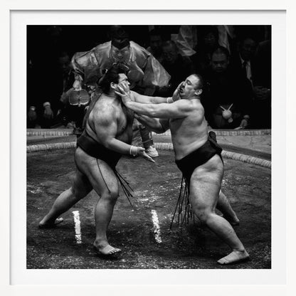 A dynamic black and white action photograph of a sumo wrestling match. Two large wrestlers are in the middle of a clay ring, one pushing forcefully against the other's face. A referee in traditional robes stands behind them, and spectators watch from the background. The entire scene is captured within a light wood frame. Artwork