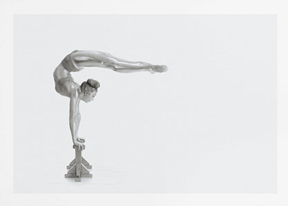 A framed black and white photograph of a female gymnast with a metallic silver-painted body, balancing in a difficult handstand pose on small blocks against a stark white background. Her back is arched and her legs are extended straight out behind her, parallel to the ground. Artwork