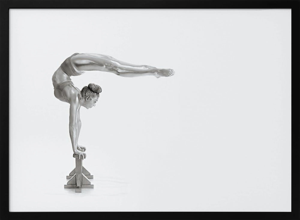 A framed black and white photograph of a female gymnast with a metallic silver-painted body, balancing in a difficult handstand pose on small blocks against a stark white background. Her back is arched and her legs are extended straight out behind her, parallel to the ground. Artwork