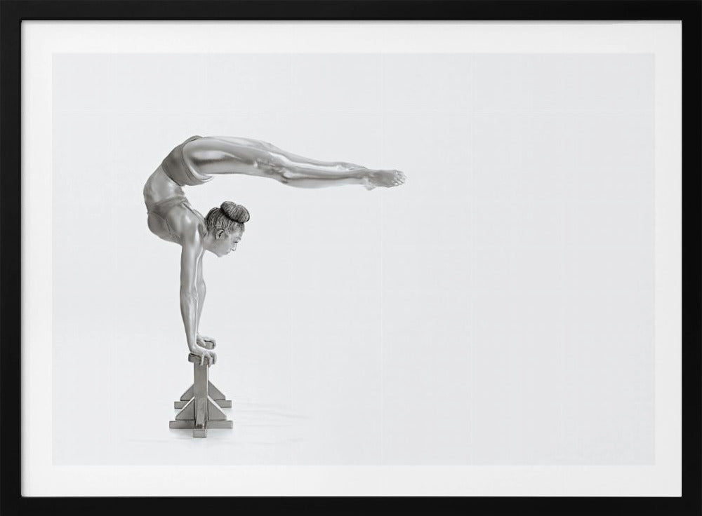 A framed black and white photograph of a female gymnast with a metallic silver-painted body, balancing in a difficult handstand pose on small blocks against a stark white background. Her back is arched and her legs are extended straight out behind her, parallel to the ground. Artwork
