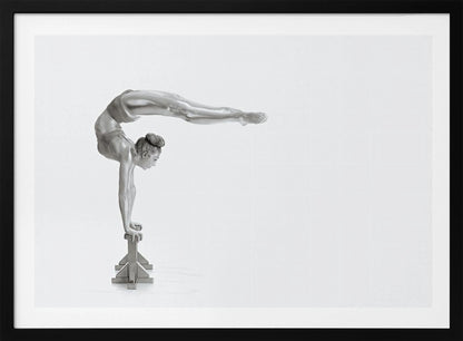 A framed black and white photograph of a female gymnast with a metallic silver-painted body, balancing in a difficult handstand pose on small blocks against a stark white background. Her back is arched and her legs are extended straight out behind her, parallel to the ground. Artwork