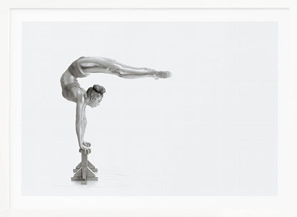 A framed black and white photograph of a female gymnast with a metallic silver-painted body, balancing in a difficult handstand pose on small blocks against a stark white background. Her back is arched and her legs are extended straight out behind her, parallel to the ground. Artwork