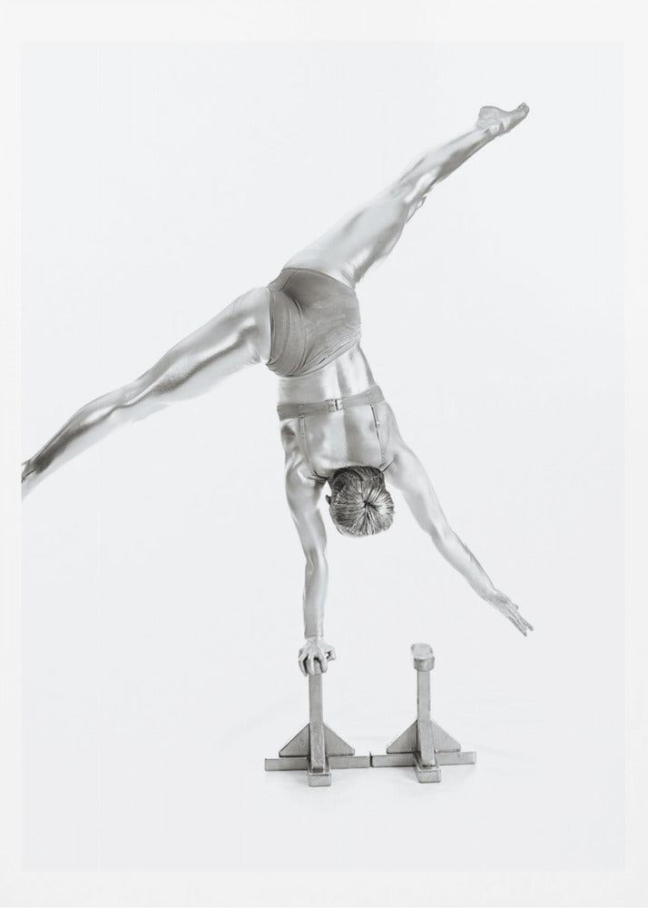 A black and white studio photograph of a muscular gymnast coated in silver paint, performing a dynamic one-armed handstand on a small block. Their legs are split wide apart in a display of strength and flexibility against a clean white background, all enclosed in a black frame. Decor