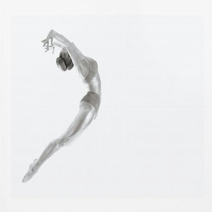 A minimalist black and white photograph of a graceful dancer frozen in mid-air, her body arched backward in an elegant C-shape against a stark white background. The artwork is presented in a light wood frame. Decor