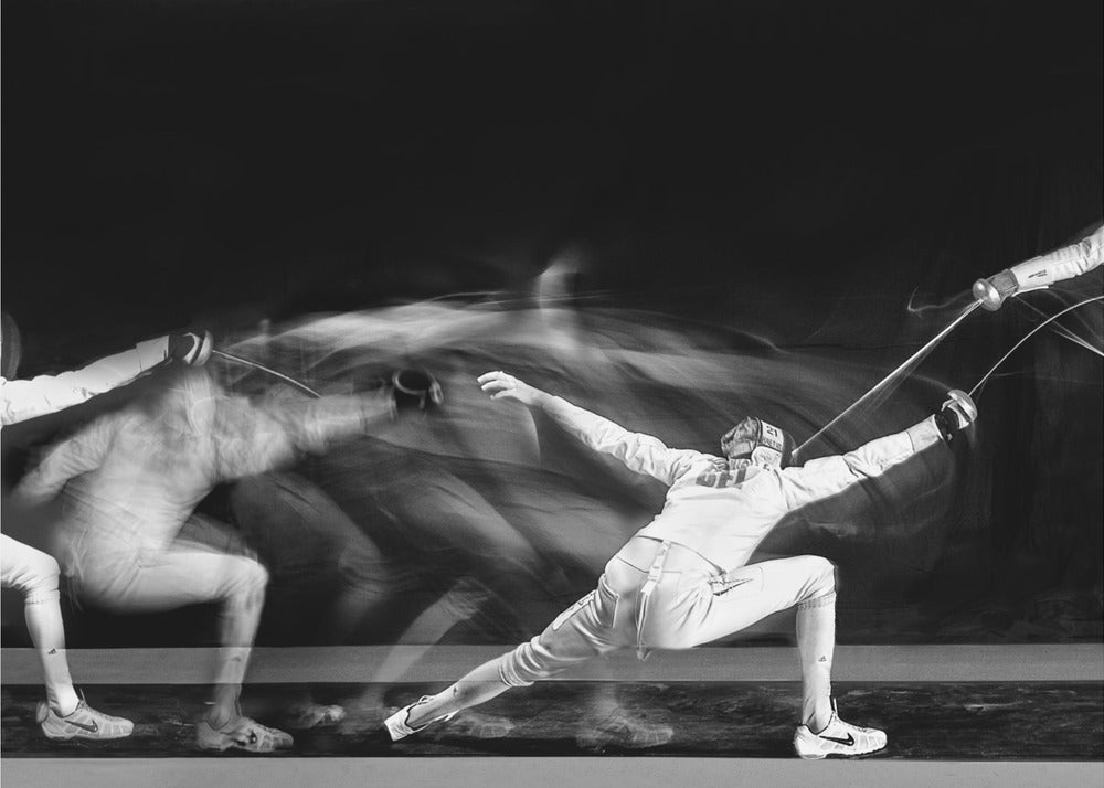 A framed, dramatic black and white photograph using long exposure to capture a fencer in motion. The fencer, in full white gear, lunges forward, their form crisp at the end of the movement while a ghostly blur trails behind, illustrating their rapid advance against a black background. Decor