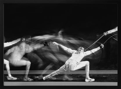 A framed, dramatic black and white photograph using long exposure to capture a fencer in motion. The fencer, in full white gear, lunges forward, their form crisp at the end of the movement while a ghostly blur trails behind, illustrating their rapid advance against a black background. Decor