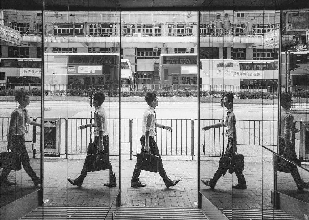 A black and white street photograph showing a man in a shirt and tie carrying a briefcase, his walking motion captured in a time-lapse effect across multiple reflective glass panels. The reflections create a sequence of his stride, set against the backdrop of a busy city street with buses and buildings also reflected in the glass. Decor