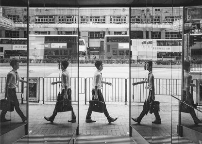 A black and white street photograph showing a man in a shirt and tie carrying a briefcase, his walking motion captured in a time-lapse effect across multiple reflective glass panels. The reflections create a sequence of his stride, set against the backdrop of a busy city street with buses and buildings also reflected in the glass. Decor