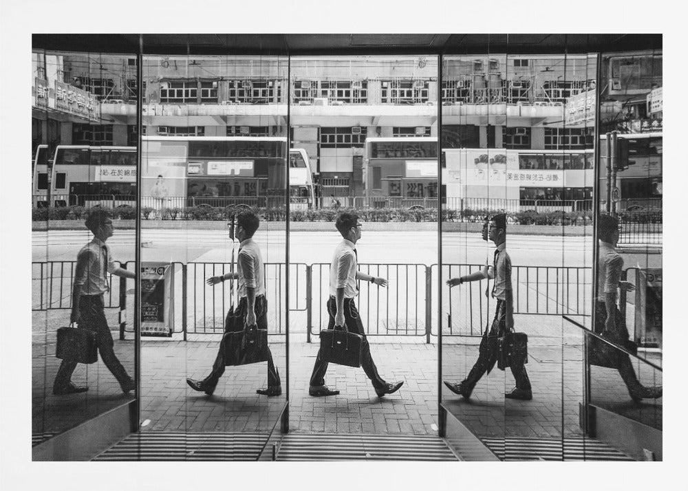 A black and white street photograph showing a man in a shirt and tie carrying a briefcase, his walking motion captured in a time-lapse effect across multiple reflective glass panels. The reflections create a sequence of his stride, set against the backdrop of a busy city street with buses and buildings also reflected in the glass. Decor