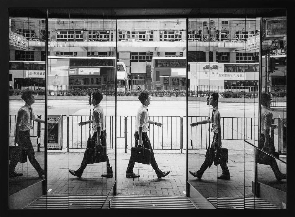 A black and white street photograph showing a man in a shirt and tie carrying a briefcase, his walking motion captured in a time-lapse effect across multiple reflective glass panels. The reflections create a sequence of his stride, set against the backdrop of a busy city street with buses and buildings also reflected in the glass. Decor