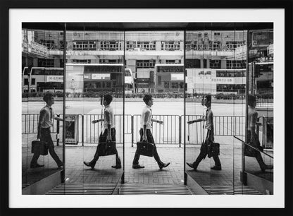 A black and white street photograph showing a man in a shirt and tie carrying a briefcase, his walking motion captured in a time-lapse effect across multiple reflective glass panels. The reflections create a sequence of his stride, set against the backdrop of a busy city street with buses and buildings also reflected in the glass. Decor