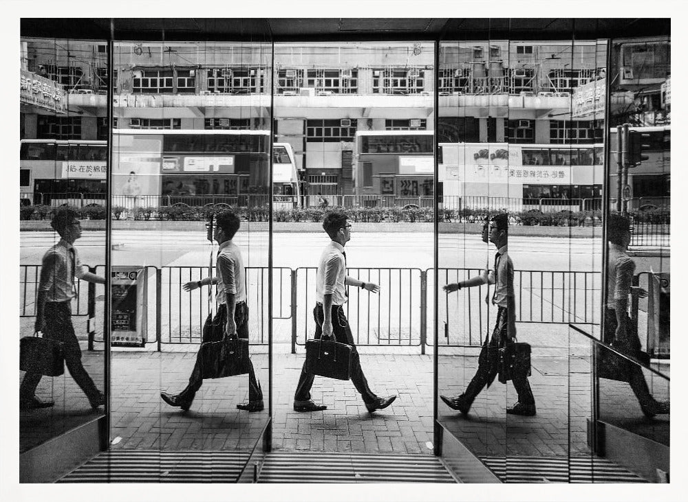A black and white street photograph showing a man in a shirt and tie carrying a briefcase, his walking motion captured in a time-lapse effect across multiple reflective glass panels. The reflections create a sequence of his stride, set against the backdrop of a busy city street with buses and buildings also reflected in the glass. Decor