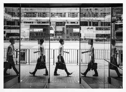 A black and white street photograph showing a man in a shirt and tie carrying a briefcase, his walking motion captured in a time-lapse effect across multiple reflective glass panels. The reflections create a sequence of his stride, set against the backdrop of a busy city street with buses and buildings also reflected in the glass. Decor