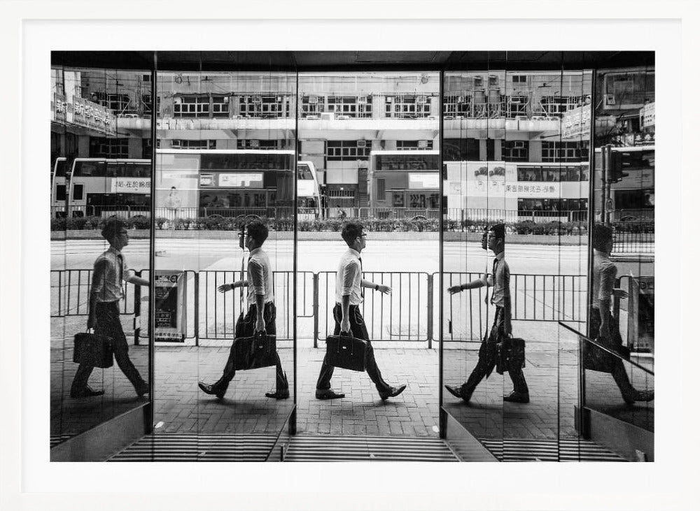 A black and white street photograph showing a man in a shirt and tie carrying a briefcase, his walking motion captured in a time-lapse effect across multiple reflective glass panels. The reflections create a sequence of his stride, set against the backdrop of a busy city street with buses and buildings also reflected in the glass. Decor