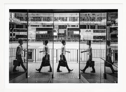 A black and white street photograph showing a man in a shirt and tie carrying a briefcase, his walking motion captured in a time-lapse effect across multiple reflective glass panels. The reflections create a sequence of his stride, set against the backdrop of a busy city street with buses and buildings also reflected in the glass. Decor