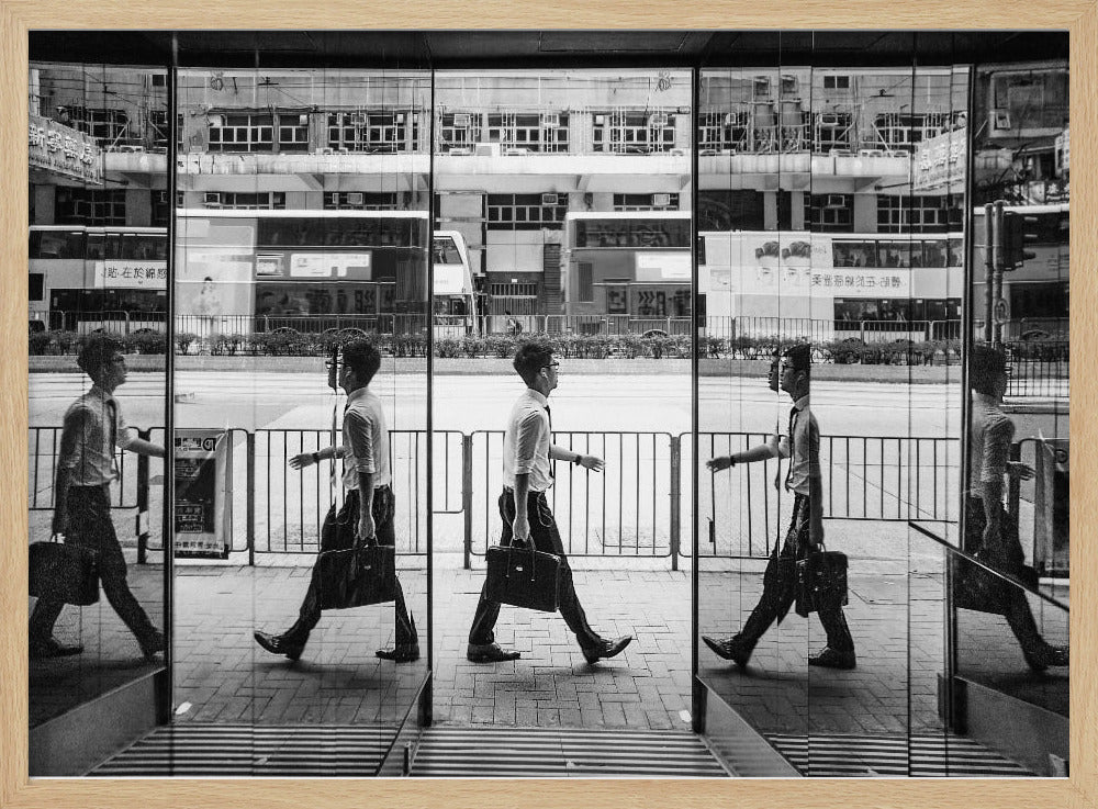 A black and white street photograph showing a man in a shirt and tie carrying a briefcase, his walking motion captured in a time-lapse effect across multiple reflective glass panels. The reflections create a sequence of his stride, set against the backdrop of a busy city street with buses and buildings also reflected in the glass. Decor