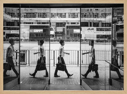 A black and white street photograph showing a man in a shirt and tie carrying a briefcase, his walking motion captured in a time-lapse effect across multiple reflective glass panels. The reflections create a sequence of his stride, set against the backdrop of a busy city street with buses and buildings also reflected in the glass. Decor