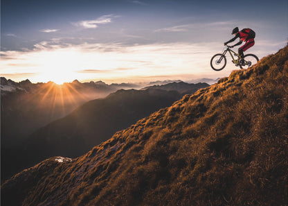 A mountain biker in mid-air, jumping down a steep grassy mountain ridge at sunrise. The sun is low on the horizon, casting a golden glow and long sunbeams over a vast, layered mountain range in the background. Poster