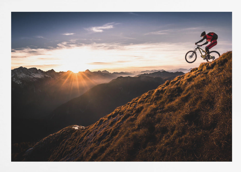 A mountain biker in mid-air, jumping down a steep grassy mountain ridge at sunrise. The sun is low on the horizon, casting a golden glow and long sunbeams over a vast, layered mountain range in the background. Poster