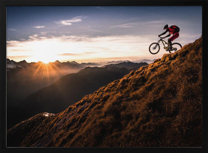 A mountain biker in mid-air, jumping down a steep grassy mountain ridge at sunrise. The sun is low on the horizon, casting a golden glow and long sunbeams over a vast, layered mountain range in the background. Poster