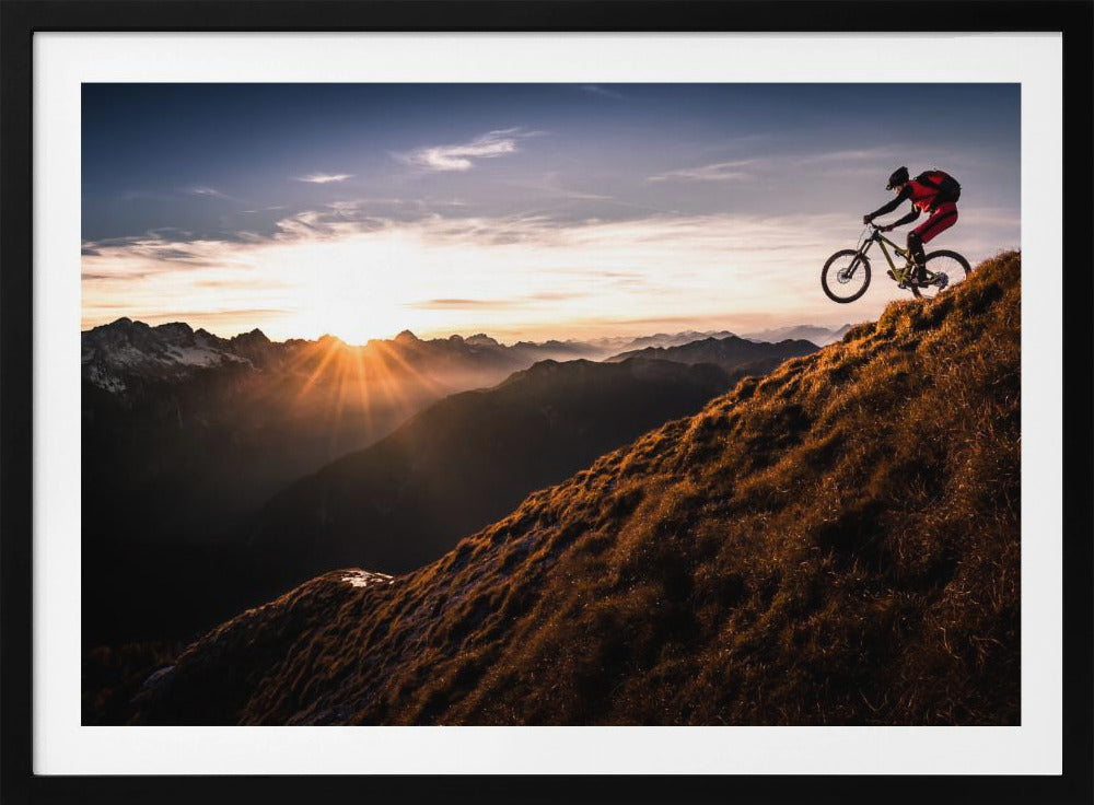 A mountain biker in mid-air, jumping down a steep grassy mountain ridge at sunrise. The sun is low on the horizon, casting a golden glow and long sunbeams over a vast, layered mountain range in the background. Poster