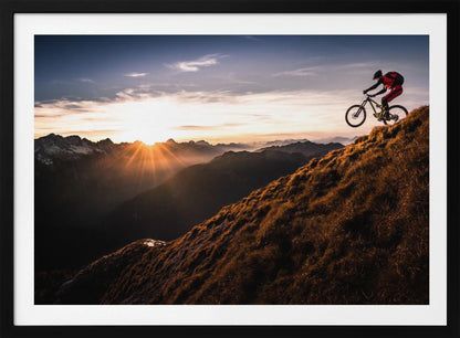 A mountain biker in mid-air, jumping down a steep grassy mountain ridge at sunrise. The sun is low on the horizon, casting a golden glow and long sunbeams over a vast, layered mountain range in the background. Poster