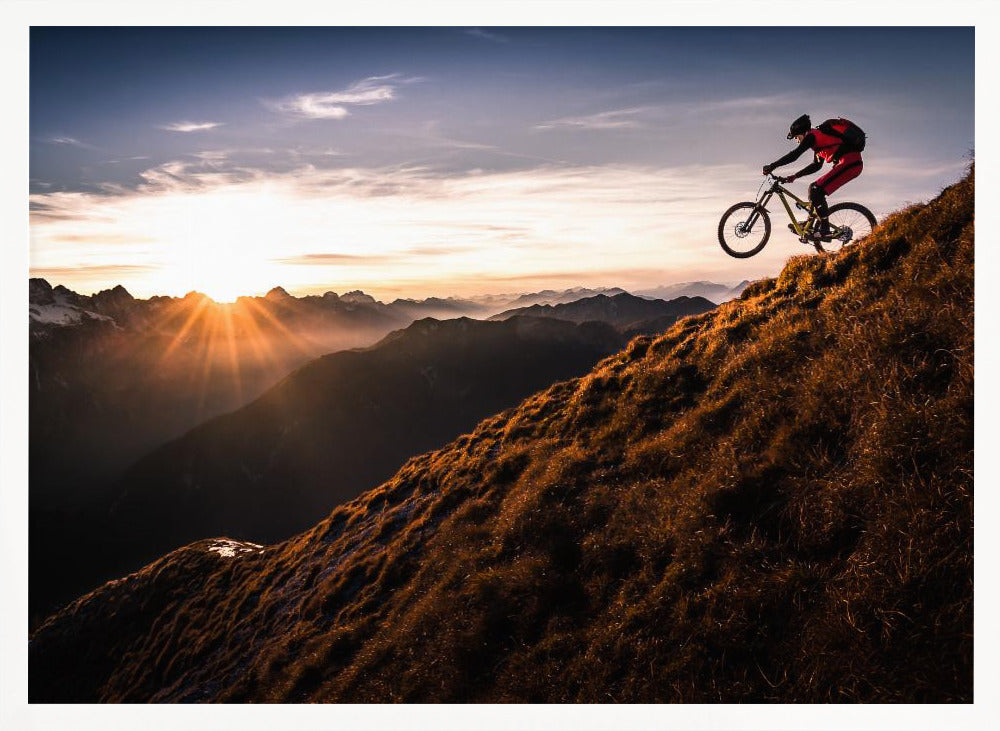 A mountain biker in mid-air, jumping down a steep grassy mountain ridge at sunrise. The sun is low on the horizon, casting a golden glow and long sunbeams over a vast, layered mountain range in the background. Poster