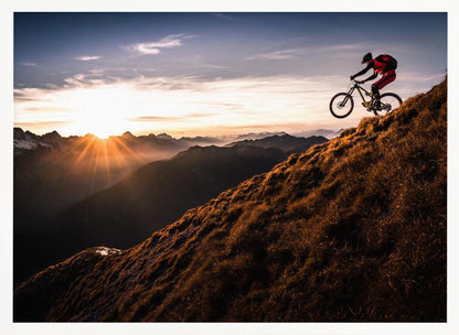A mountain biker in mid-air, jumping down a steep grassy mountain ridge at sunrise. The sun is low on the horizon, casting a golden glow and long sunbeams over a vast, layered mountain range in the background. Poster