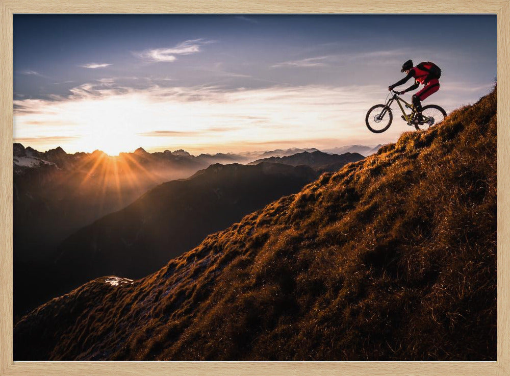 A mountain biker in mid-air, jumping down a steep grassy mountain ridge at sunrise. The sun is low on the horizon, casting a golden glow and long sunbeams over a vast, layered mountain range in the background. Poster