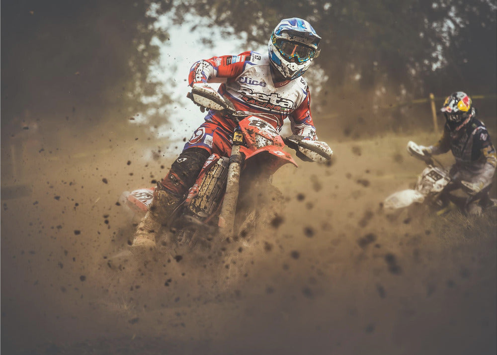 An action shot of a motocross rider in red, white, and blue gear, speeding through a dusty turn on a dirt track, kicking up a large cloud of mud and debris. Another racer is visible in the background. The image is presented within a silver frame. Print