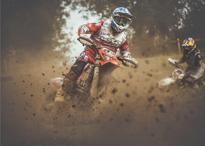 An action shot of a motocross rider in red, white, and blue gear, speeding through a dusty turn on a dirt track, kicking up a large cloud of mud and debris. Another racer is visible in the background. The image is presented within a silver frame. Print