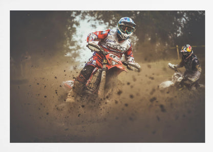 An action shot of a motocross rider in red, white, and blue gear, speeding through a dusty turn on a dirt track, kicking up a large cloud of mud and debris. Another racer is visible in the background. The image is presented within a silver frame. Print