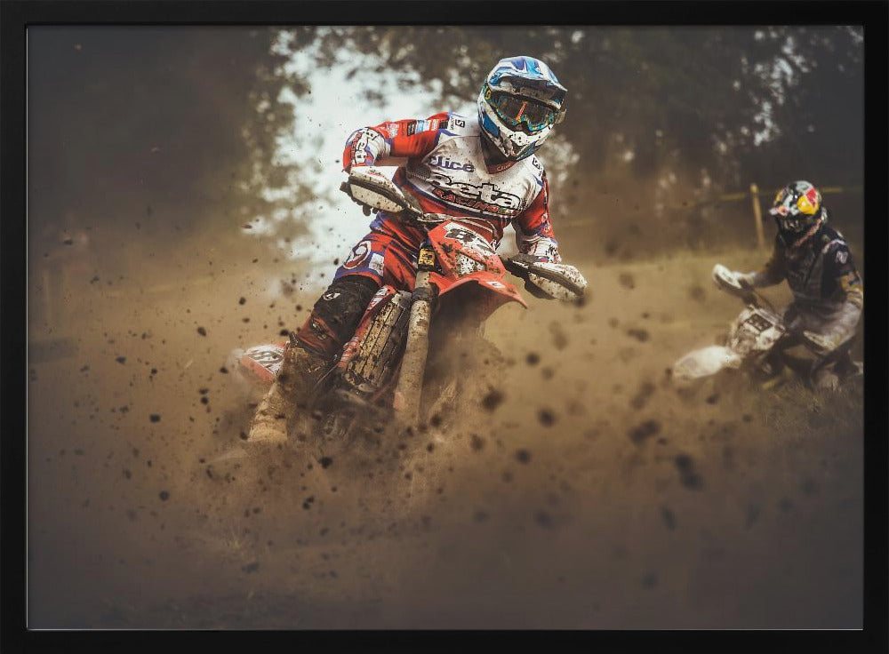 An action shot of a motocross rider in red, white, and blue gear, speeding through a dusty turn on a dirt track, kicking up a large cloud of mud and debris. Another racer is visible in the background. The image is presented within a silver frame. Print