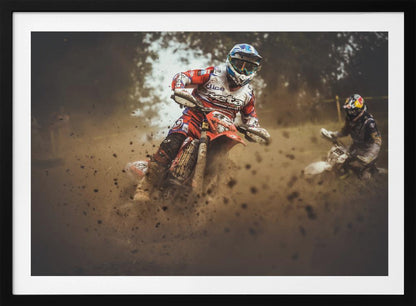 An action shot of a motocross rider in red, white, and blue gear, speeding through a dusty turn on a dirt track, kicking up a large cloud of mud and debris. Another racer is visible in the background. The image is presented within a silver frame. Print